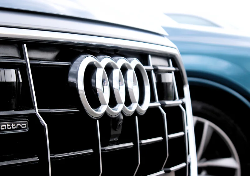 Audi logo on black grille with silver trim and neighboring blue car